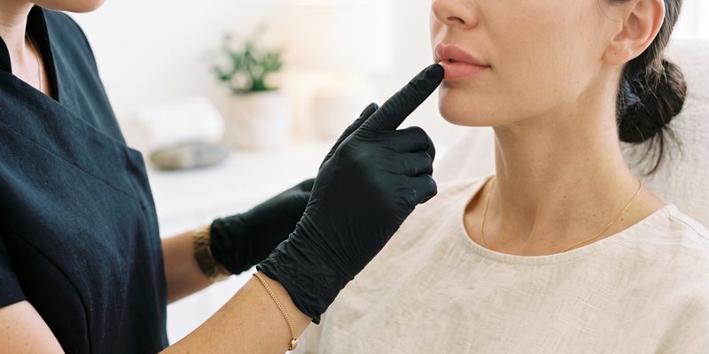 A gloved practitioner assessing a client's lips with a fingertip before treatment